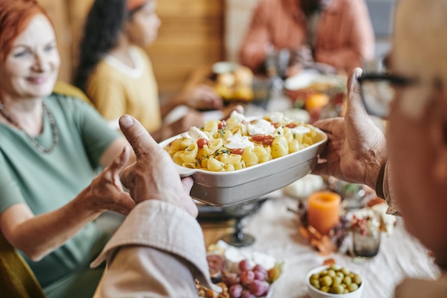 Mão de um homem maduro passando a tigela com massa assada para a esposa, enquanto os dois estão sentados à mesa festiva contra a família