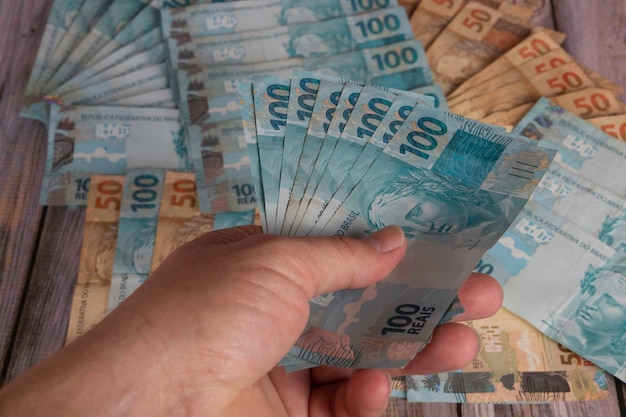 Foto mão de um homem segurando notas de 100 reais com notas de 50 e 100 reais ao fundo foco seletivo