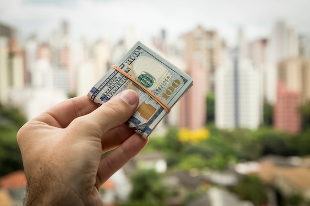 Mão de um homem segurando notas de dólar em frente a uma paisagem da cidade