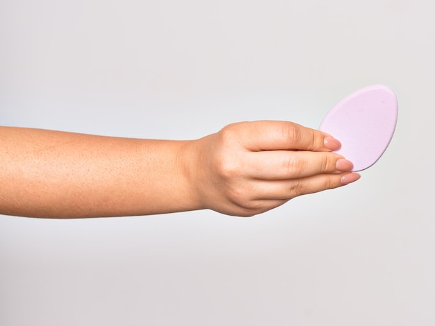 Mão de uma jovem caucasiana segurando uma esponja de maquiagem rosa sobre fundo branco isolado