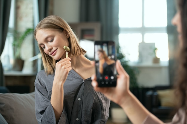 Mão de uma jovem com smartphone atirando em sua amiga usando o rolo de massagem no rosto e apreciando o procedimento de beleza