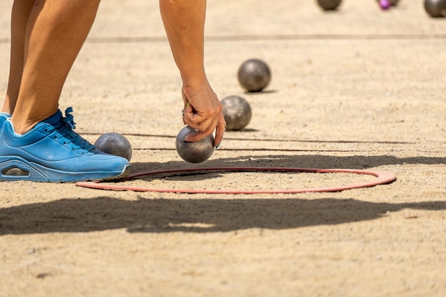Mão de uma jovem esportiva em calçados esportivos pegando uma bola de petanca de metal para competir em um terreno arenoso
