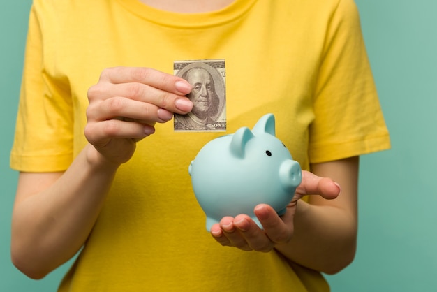 Mão de uma mulher colocando 100 dólares no porquinho para economizar dinheiro, riqueza e conceito financeiro.