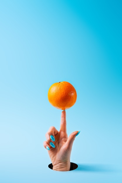 Mão de uma mulher com unhas pintadas, equilibrando a fruta laranja no dedo indicador através de um buraco cortado em papel azul