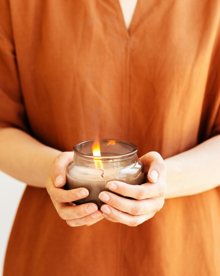 Foto mão de uma mulher segura uma vela perfumada acesa em um frasco de vidro com ingredientes naturais em linho de terracota