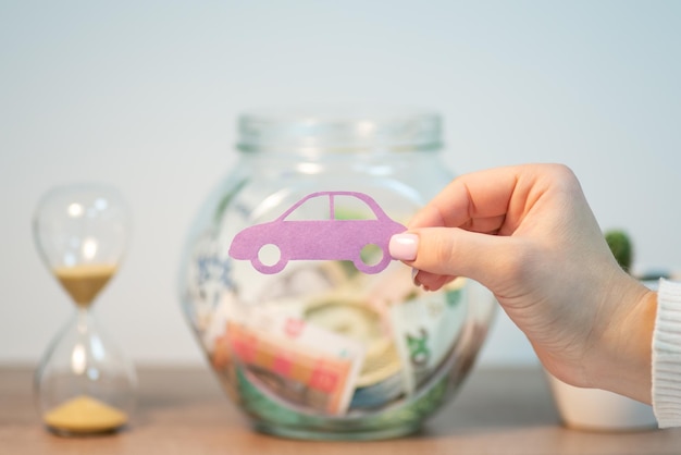 Mão de uma mulher segurando a foto da jarra de vidro do carro com dinheiro e ampulheta sobre a mesa no fundo Conceito de economizar dinheiro investindo no planejamento financeiro do veículo