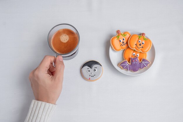 Mão de uma mulher segurando a xícara de café durante a ingestão de biscoitos de Halloween engraçados. Feliz Dia das Bruxas, Truque ou Ameaça, Olá outubro, outono outono, Tradicional, conceito de festa e feriado