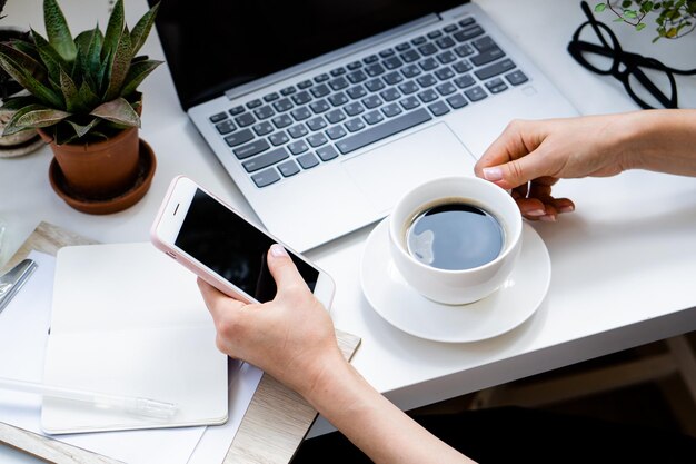 Mão de uma mulher segurando a xícara de café e smartphone no escritório aconchegante com laptop e plantas verdes