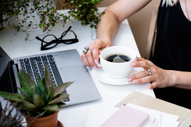 Mão de uma mulher segurando a xícara de café no aconchegante escritório de verão com laptop e plantas verdes