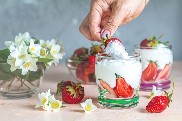 Mão de uma mulher segurando creme de morango Tigela de vidro de morangos com chantilly e hortelã Flores de jasmim Espaço de cópia de superfície de concreto para você texto