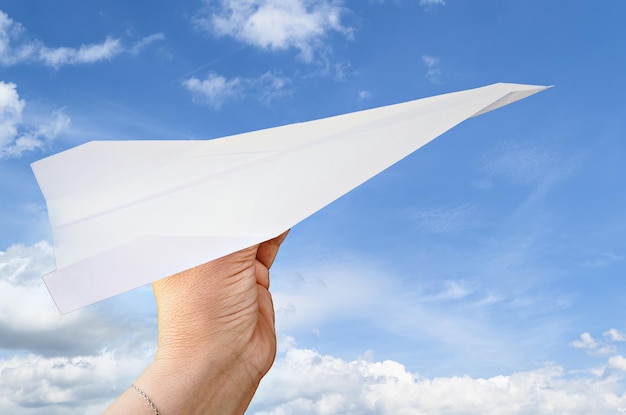Mão de uma mulher segurando um avião de papel no fundo do céu azul com nuvens brancas