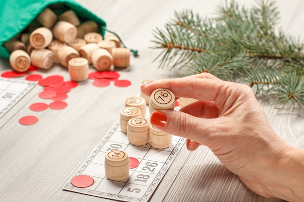 Mão de uma mulher segurando um barril para um jogo de loteria. Barris de loto de madeira com saco verde, cartas de jogo e fichas vermelhas, galhos de árvores de abeto de Natal no fundo. Loto de jogo de tabuleiro