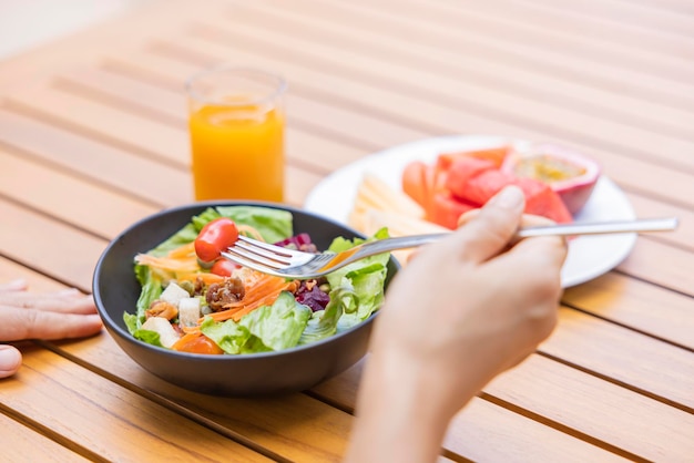 Mão de uma mulher segurando um garfo e comendo café da manhã Frutas de salada de vegetais como melancia mamão melão maracujá suco de laranja e café colocado em um jogo americano cinza