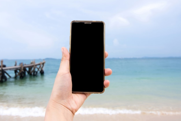 Mão de uma mulher segurando um smartphone com fundo de praia do mar com traçado de recorte na tela de exibição