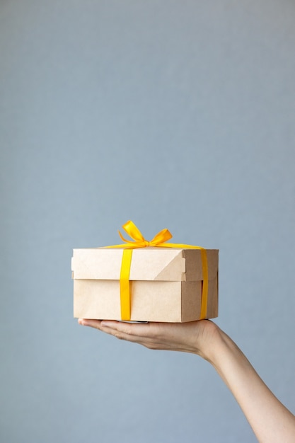 Mão de uma mulher segurando uma caixa de papelão kraft de cor natural. Mulher segurando uma caixa com uma fita amarela de presente