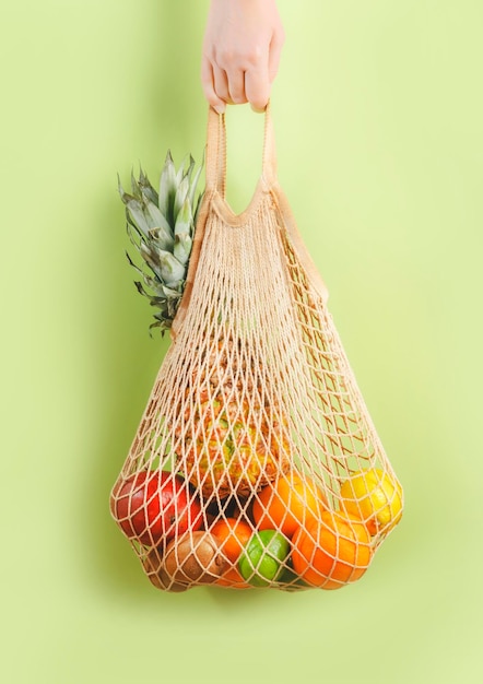 Mão de uma mulher segurando uma sacola ecológica com várias frutas tropicais em fundo verde Estilo de vida sustentável Sem plástico Conceito de desperdício zero