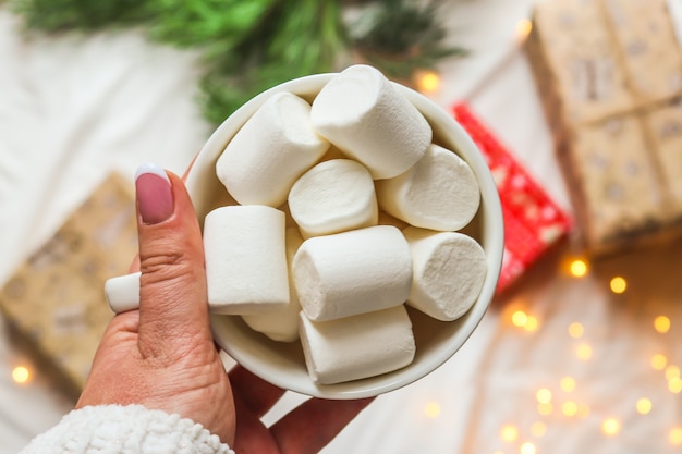 Mão de uma mulher segurando uma xícara com marshmallows no fundo desfocado de feriado. Festivo e feriado