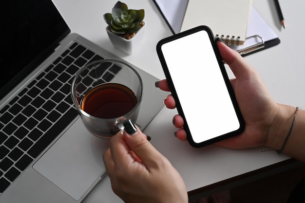 Mão de uma mulher segurando uma xícara de café e usando o celular na mesa de escritório