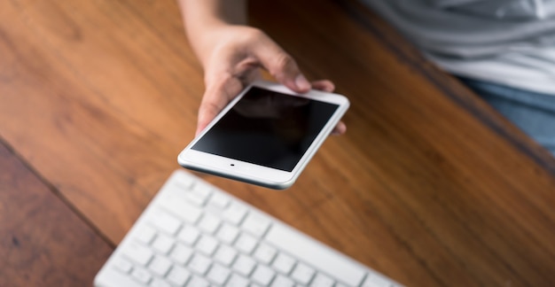 Mão de uma mulher usando telefone inteligente com a área de trabalho do computador na mesa de madeira