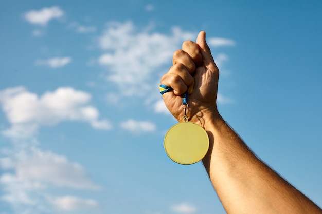 Foto mão de vencedor levantada e segurando a medalha de ouro com fita contra o céu azul.