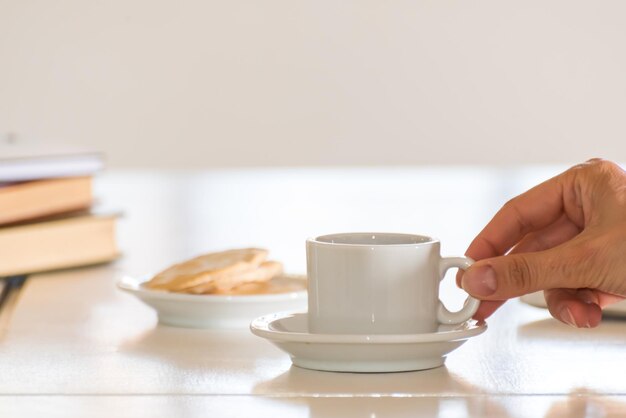 Mão delicada segurando uma xícara de café branca com livros fora de foco atrás dela