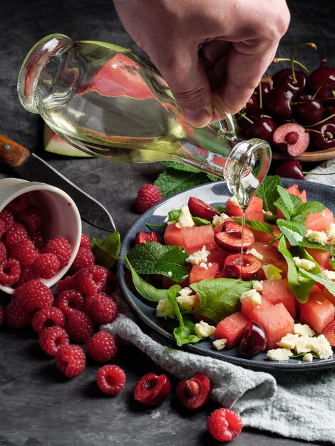 Mão derramando óleo sobre a salada com melancia, queijo, hortelã, frutas vermelhas e cerejas no fundo cinza