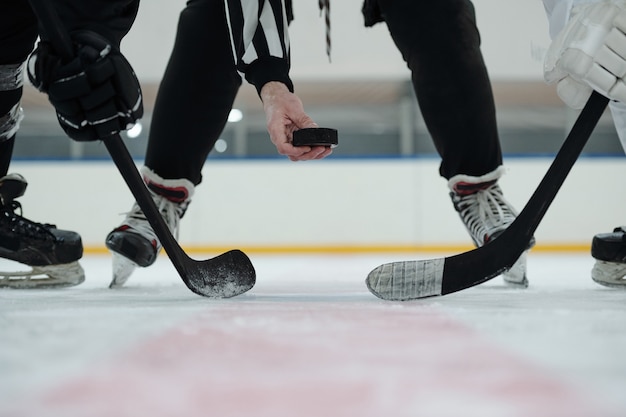 Foto mão do árbitro segurando o disco sobre a pista de gelo com dois jogadores com tacos parados e esperando o momento de atirar