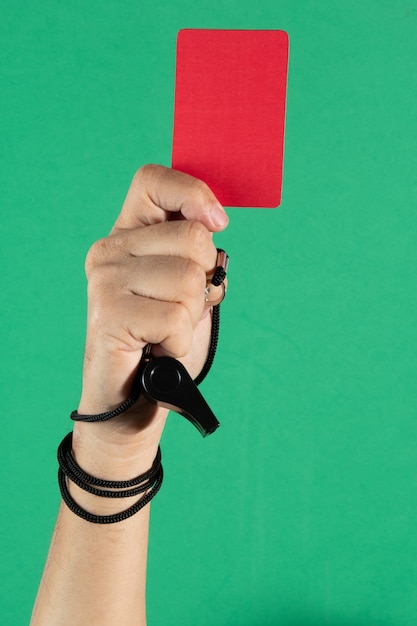 Mão do árbitro segurando um cartão vermelho e apito sobre fundo verde.