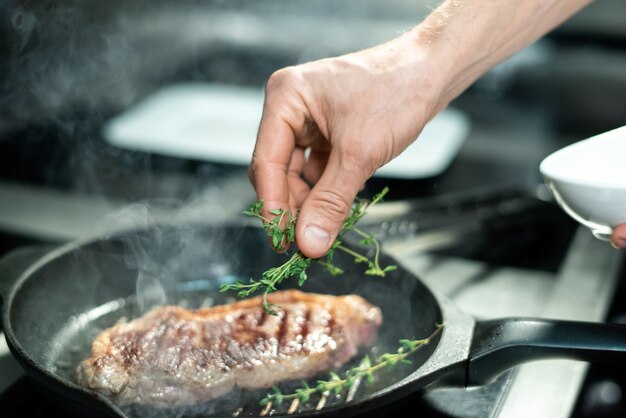 Mão do chef colocando ervas aromáticas verdes em um pedaço de carne assada na frigideira quente enquanto fica perto do fogão elétrico e cozinha a carne