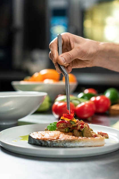 Mão do chef colocando flor de açafrão em cima de um apetitoso guisado de vegetais no prato com um pedaço de salmão assado enquanto prepara a refeição para o cliente