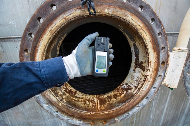 Mão do trabalhador segurando detector de gás, inspeção e teste de segurança de gás no tanque de aço inoxidável frontal para trabalhar dentro de confinamento