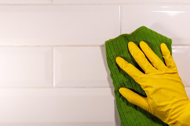 Mão em uma luva de borracha amarela, limpando a superfície do ladrilho de cerâmica com um pano verde, close-up, copie o espaço. Desinfecção da cozinha, banheiro. Conceito de limpeza de casa
