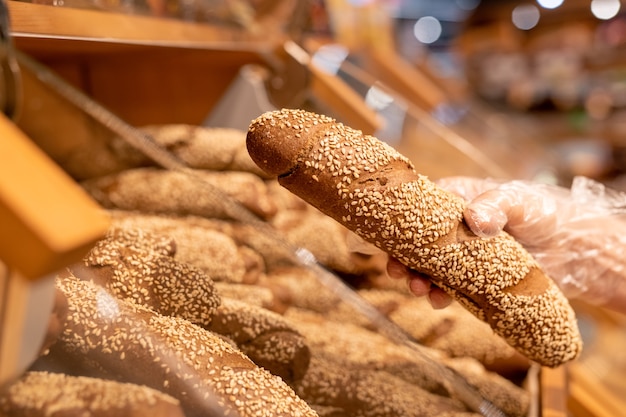 Mão enluvada de uma compradora madura segurando pão fresco polvilhado com sementes de gergelim enquanto estava em um display no supermercado