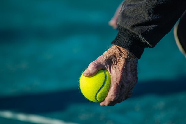 Mão envelhecida segurando uma bola de tênis na quadra