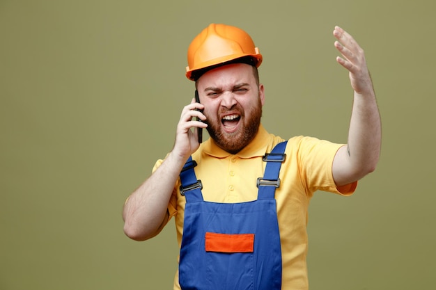 Mão espalhada animada fala no telefone jovem construtor de uniforme isolado em fundo verde