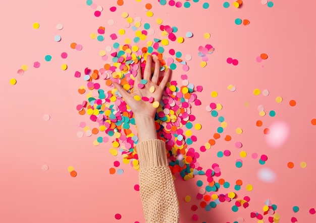 Mão estendida entre confetes coloridos em um fundo rosa simbolizando celebração e alegria