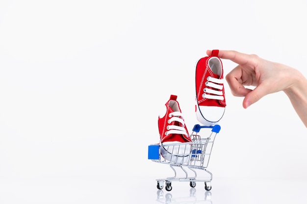 Mão feminina coloca o tênis bebê vermelho no carrinho de compras isolado no fundo branco. Loja online de calçados infantis. Copie o espaço.