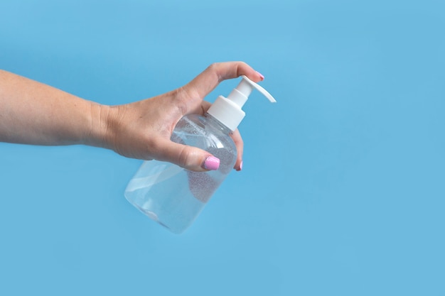 Mão feminina com manicure rosa segura a garrafa de desinfetante para as mãos transparente sobre fundo azul. Higiene das mãos. Proteção de bactérias