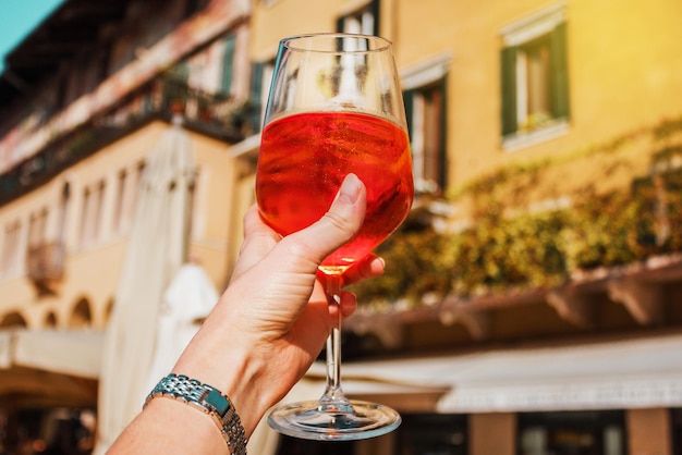 Mão feminina com um copo de spritz de coquetel laranja perto de edifícios antigos dia ensolarado em Verona, Itália