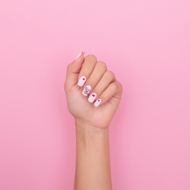 Mão feminina com unhas de manicure criativas, esmalte de gel branco, desenho de corações, em fundo rosa