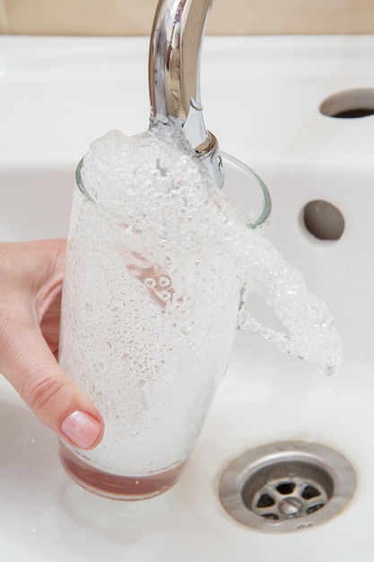 Mão feminina com vidro. O copo é enchido com água transparente sob a torneira com um lavatório de cerâmica no fundo