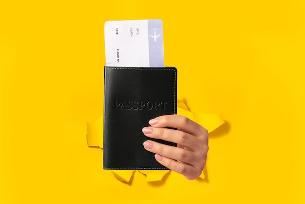 Mão feminina de turista segurando passaporte com passagem de avião isolada através de fundo amarelo rasgado