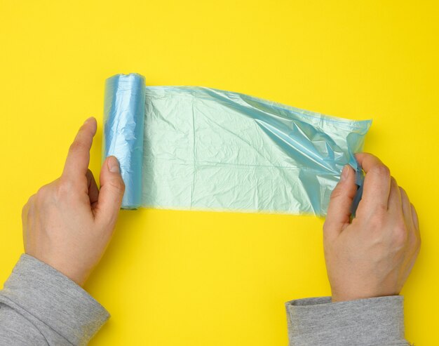 Mão feminina desenrola um saco plástico azul para lixo, superfície amarela, vista de cima