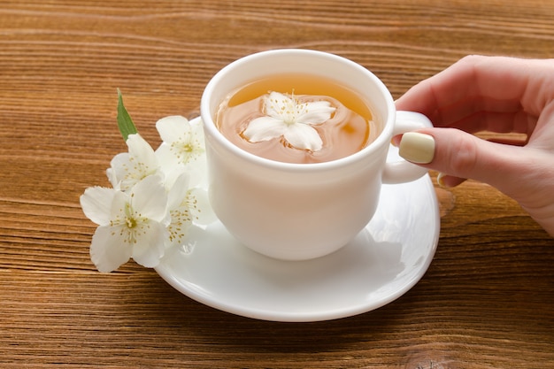 Mão feminina e uma caneca branca de chá com jasmim em uma mesa de madeira