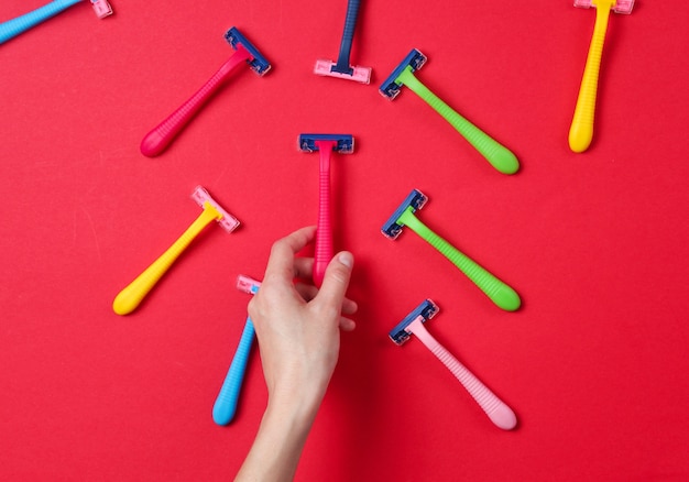 Mão feminina escolhendo navalha verde entre uma variedade de lâminas de depilação coloridas no vermelho. Vista do topo. Conceito de beleza minimalista