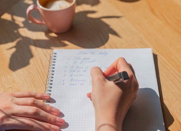 Mão feminina escrevendo planos em um caderno ou planejador sobre uma mesa de madeira com luz do sol e xícara de café