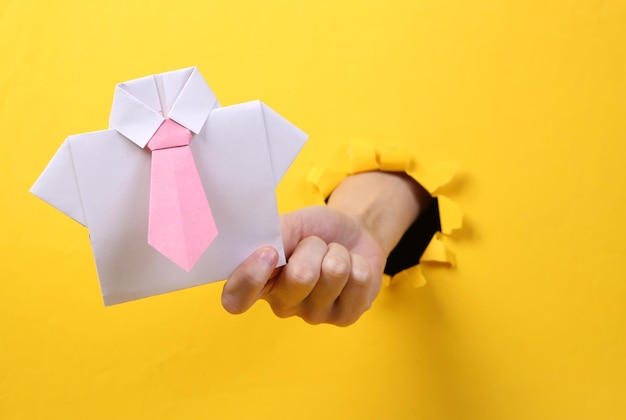 Mão feminina segura camisa de origami com gravata através de papel amarelo rasgado arte conceitual minimalismo