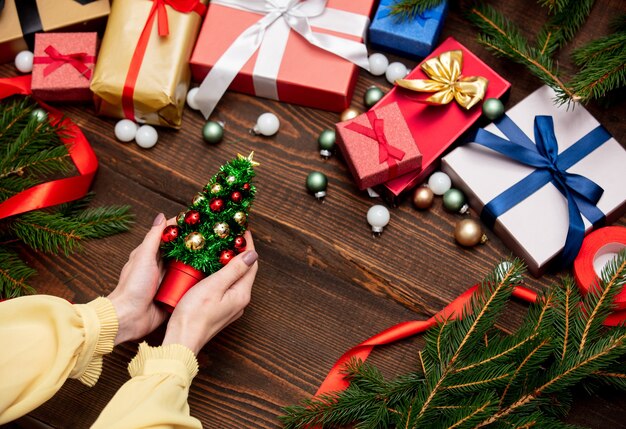 Mão feminina segura uma árvore de natal perto de presentes de natal e ramos de pinheiro, enfeites na mesa de madeira