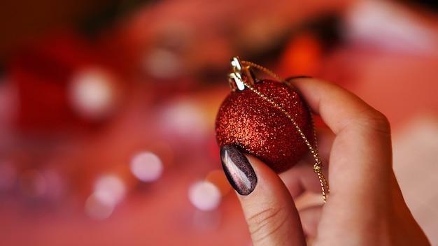 Mão feminina segura uma bola vermelha no fundo de Natal decorado - luzes de bokeh