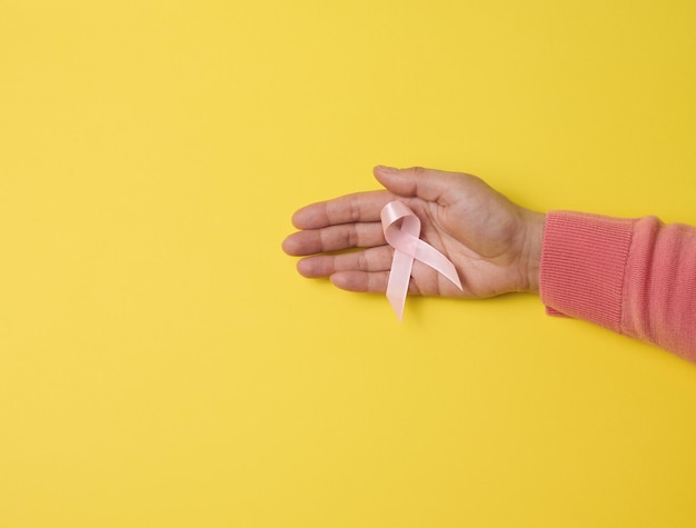 Mão feminina segura uma fita de seda rosa na forma de um laço sobre um fundo amarelo. Símbolo da luta contra o câncer de mama